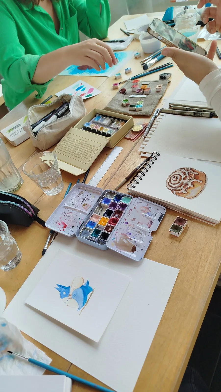 La mesa de trabajo vista a lo largo, con paletas abiertas, cuadernos y varias láminas empezadas