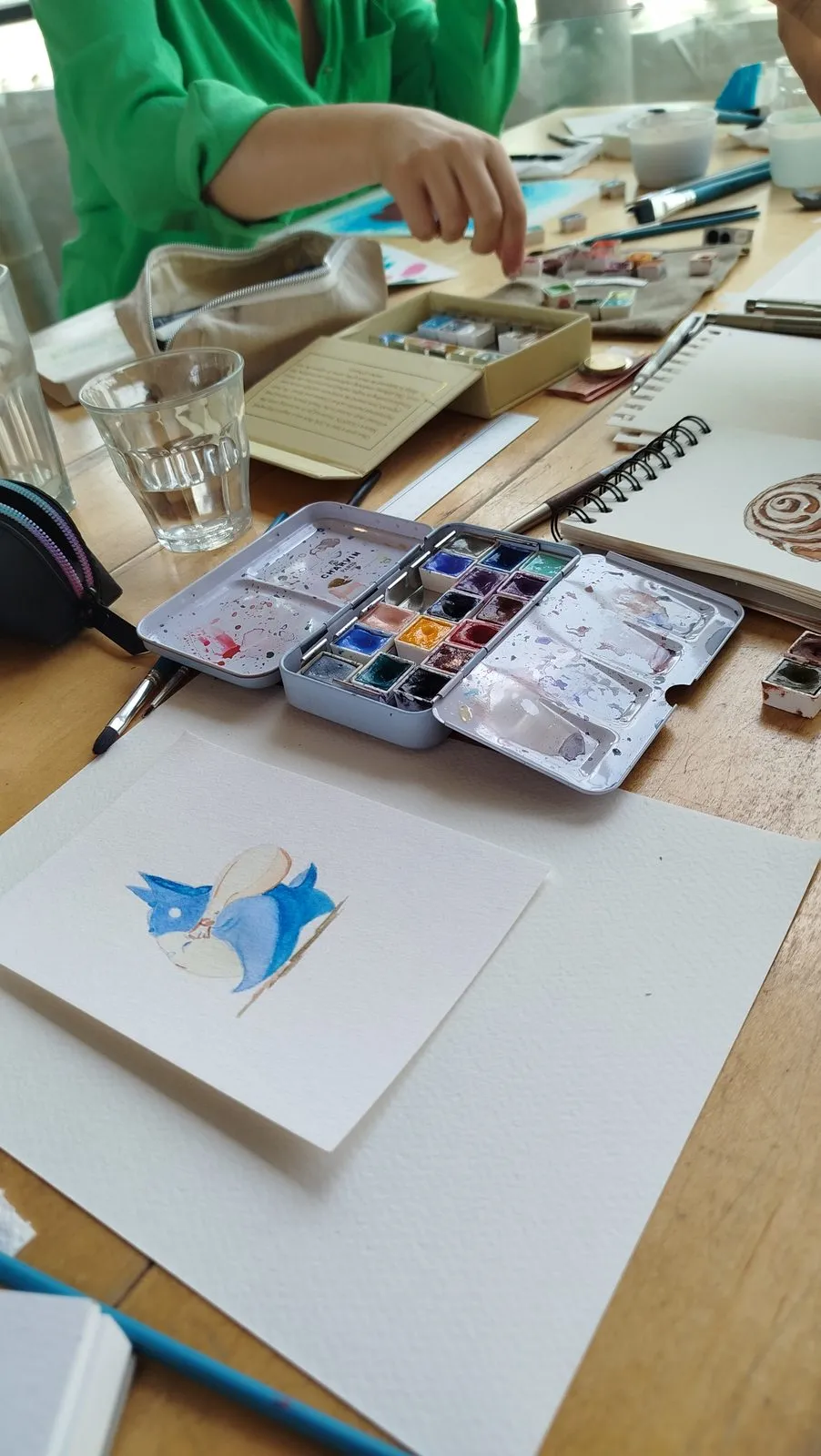 Dos personas pintan en el mismo lado de la mesa, con los pinceles y los cuadernos abiertos