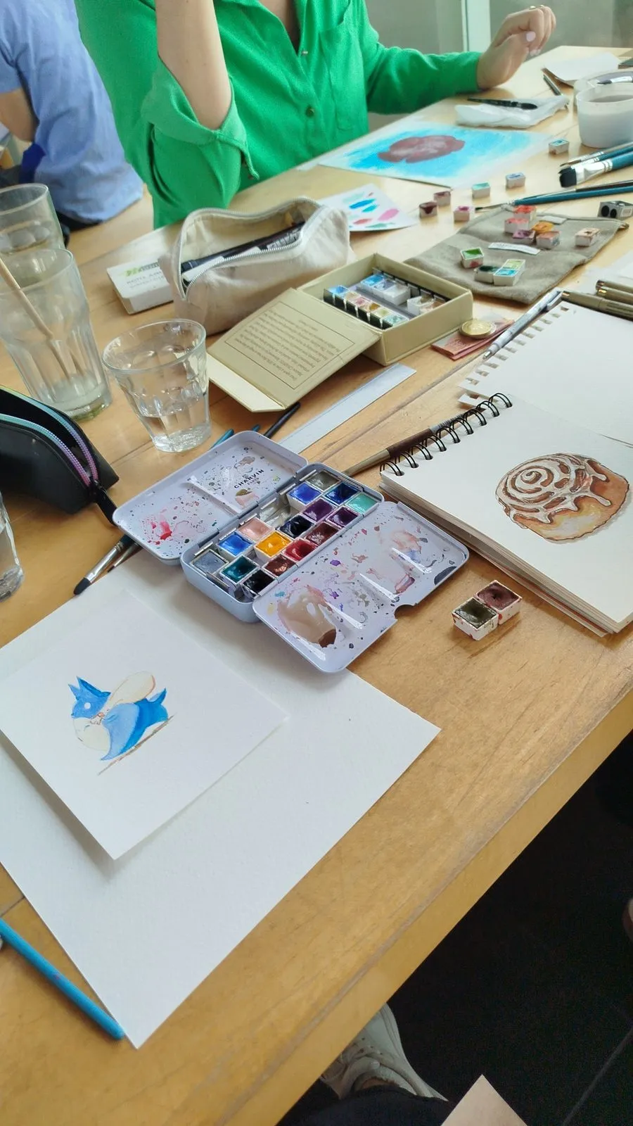 Paleta de acuarelas abierta junto a una lámina con un pájaro azul y un caracol pintado a la derecha