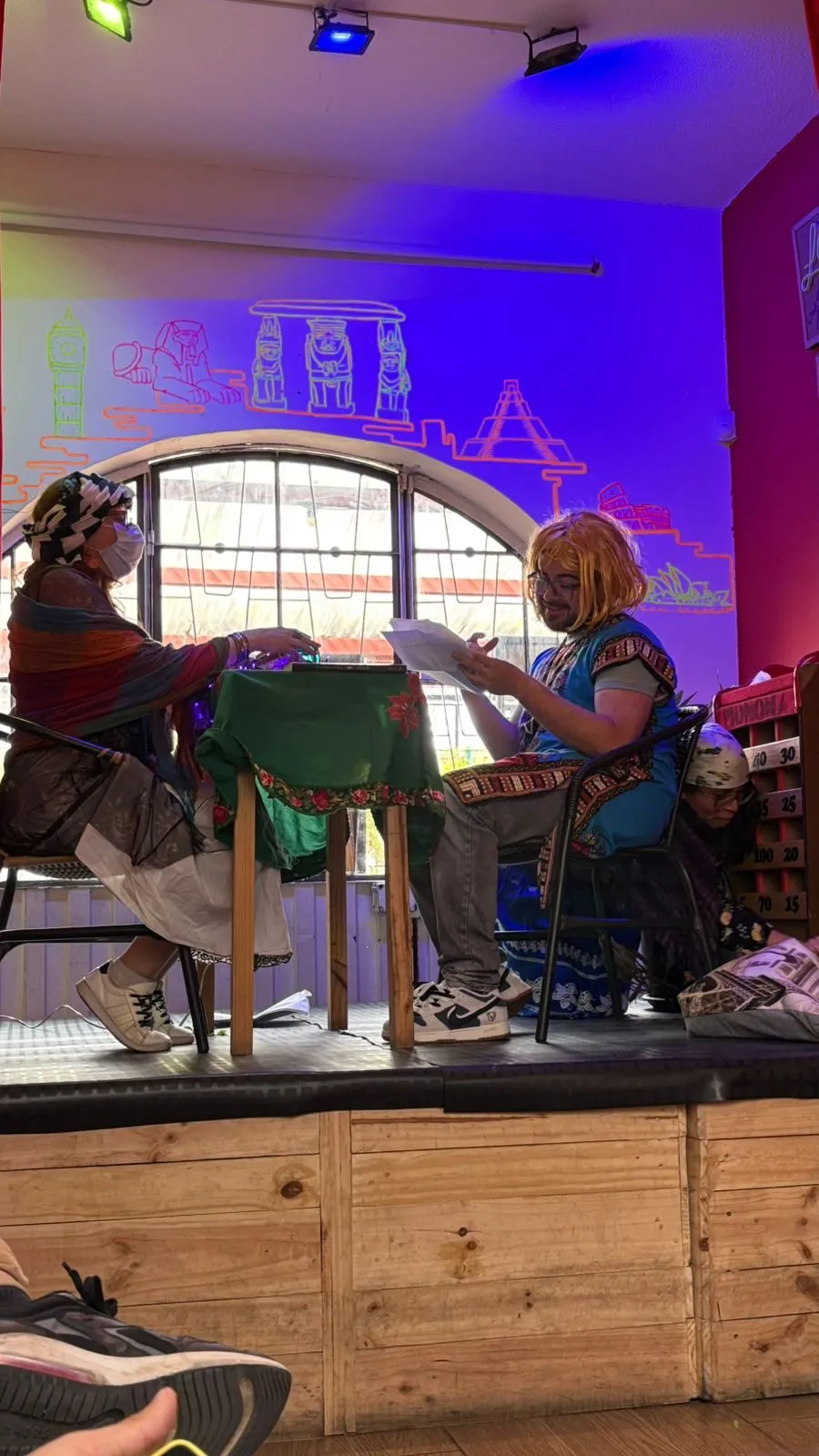Dos alumnas actúan alrededor de una mesa con mantel verde, con pelucas y vestuario